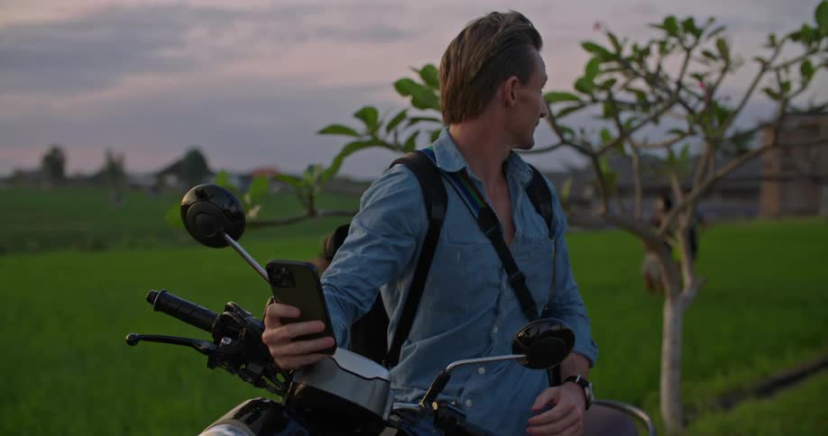 Man with mustache blue shirt and black backpack sits on moped using smartphone in the countryside