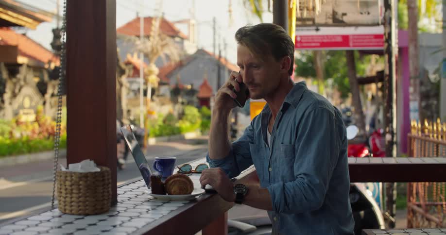 A man is sitting at an outdoor cafe with a laptop computer and talking on his cell phone while holding a coffee cup as traffic passes by on the street