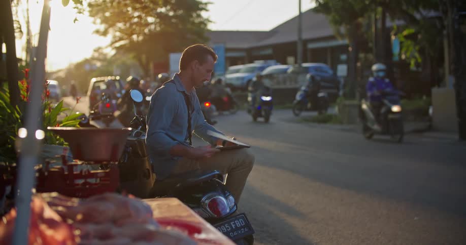 A man is sitting on his motorbike on the side of a neighborhood street typing on a laptop computer as traffic moves by on the street and he stands up to leave