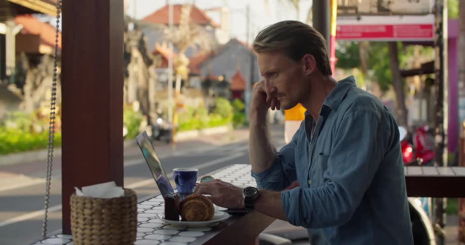 Man working on a laptop while sitting on a restaurant next to a street at evening