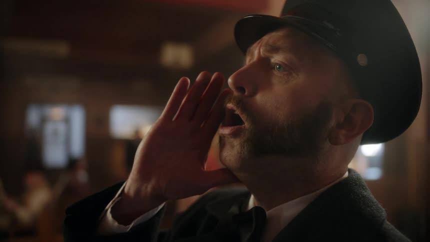 People sitting in an old time train station when a conductor calls all aboard