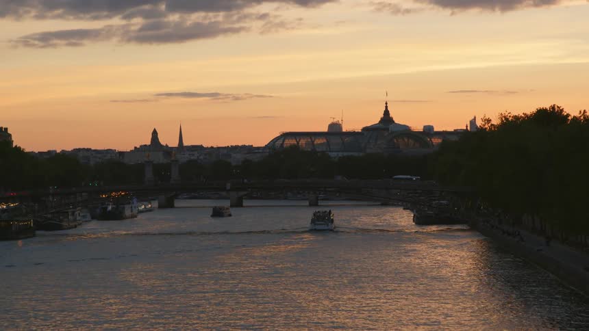 Ferries moving ahead under a bridge on a river through the silhouette of a town at evening
