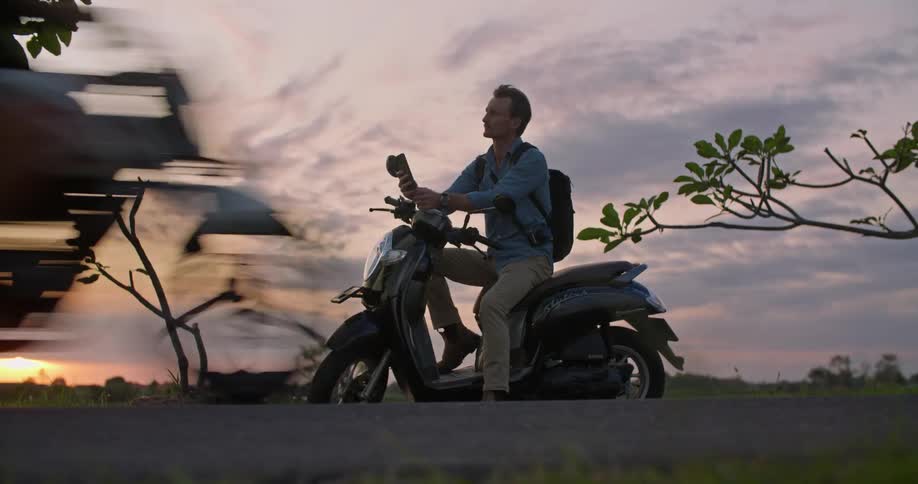 A man is outside sitting on his motor scooter and looking at his cell phone as another scooter passes on the road