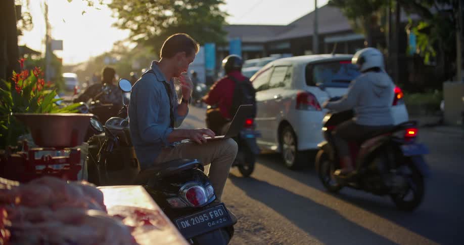 A man is sitting on his motor scooter on the side of a busy street typing on a laptop computer and he picks up his cell phone as traffic moves by on the street