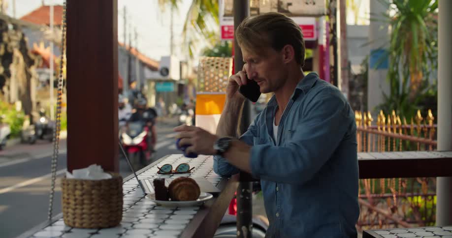A man is sitting at an outdoor cafe with a laptop computer and talking on his cell phone as motor scooters pass by on the street