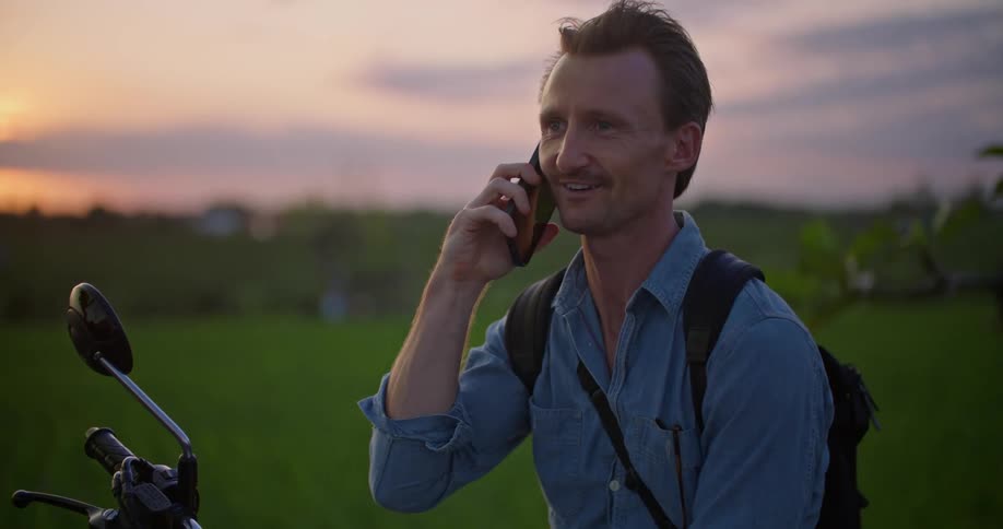 Man sitting on a parked motorcycle on a rural field talking on the phone at evening