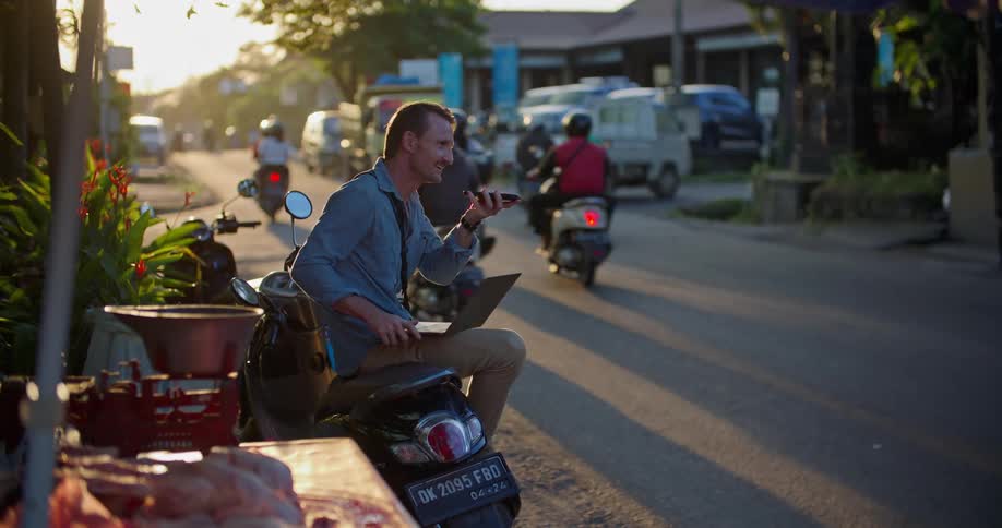 Man with mustache blue shirt and brown pants sits on moped using laptop and smartphone in the busy street