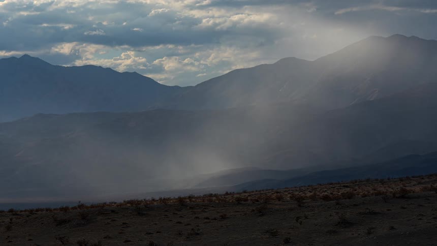 Clouds move across the sky over mountains as rays from the sun shine on the fog