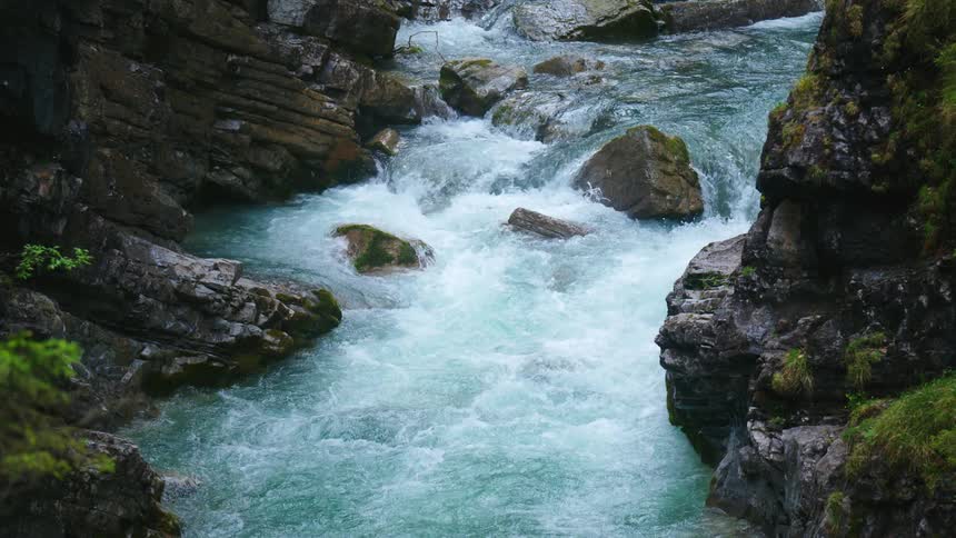 Water in a stream is rolling over and around rocks and moss covered rocks are on the riverbank