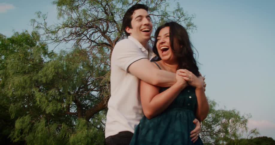 Man and woman laugh as he hugs her outdoors near the trees under a blue sky