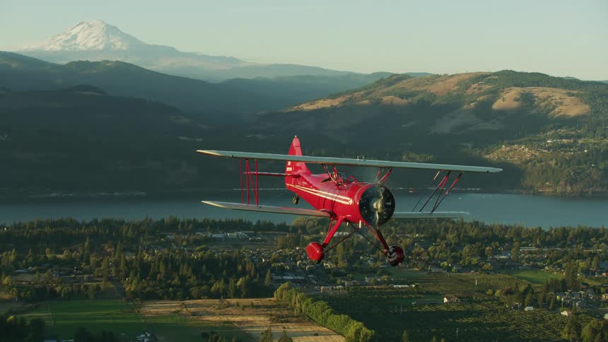 The view is of a red biplane flying over Hood River with Mount Adams in the background in Oregon
