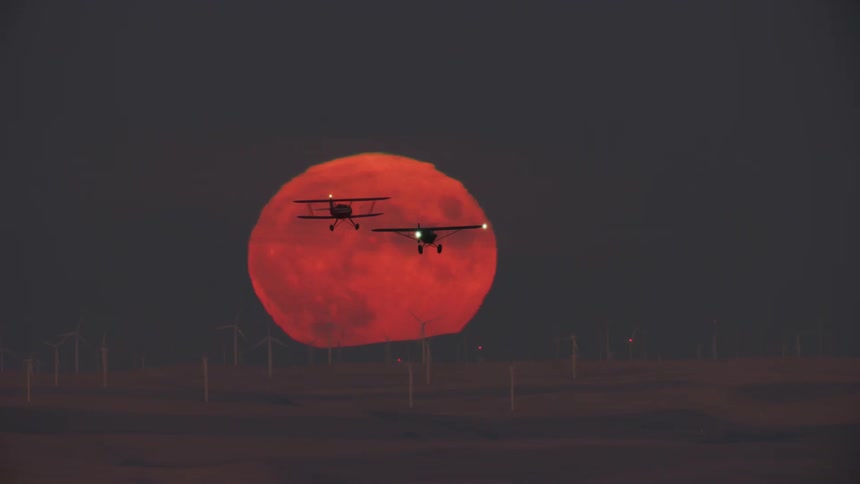 Two small airplanes fly over the wind turbines in the field as the sun sets