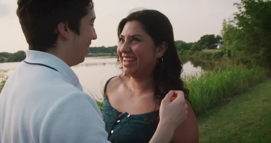 Man lovingly strokes the hair of his girlfriend as she looks at him and laughs while standing next to a lake