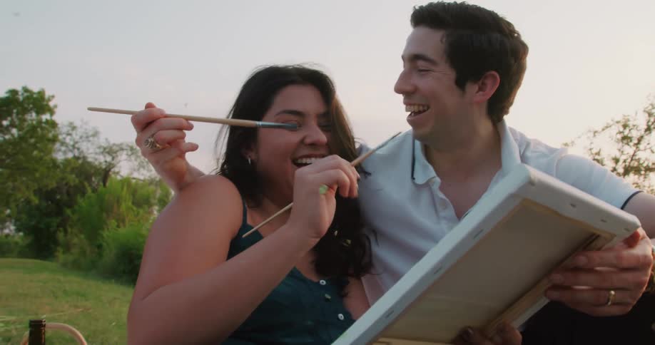 Young happy couple sitting on the grass laugh and hug while painting with oils on canvas at evening