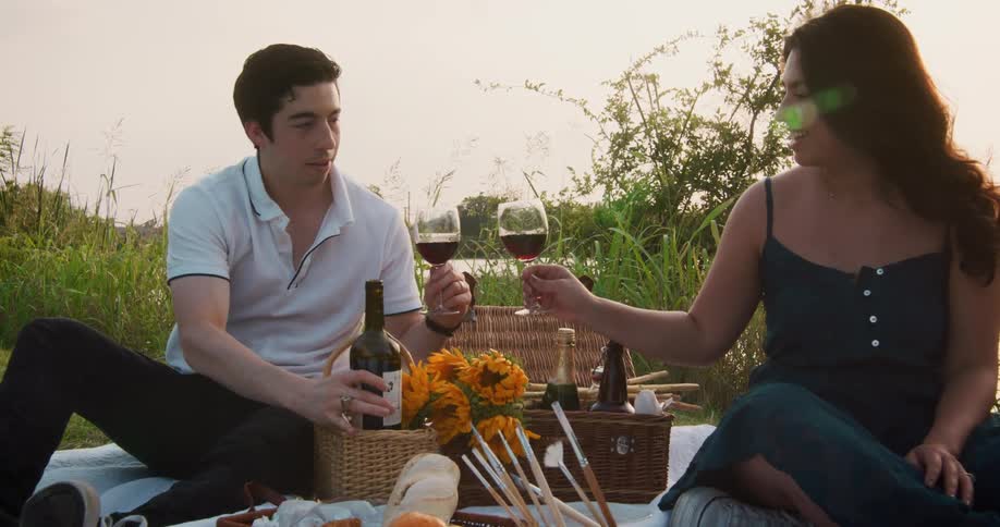 Couple drink wine while sitting on riverbank near picnic basket and artist brushes
