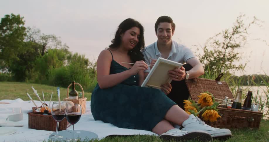 A couple is having a picnic with some wine by them and painting a picture