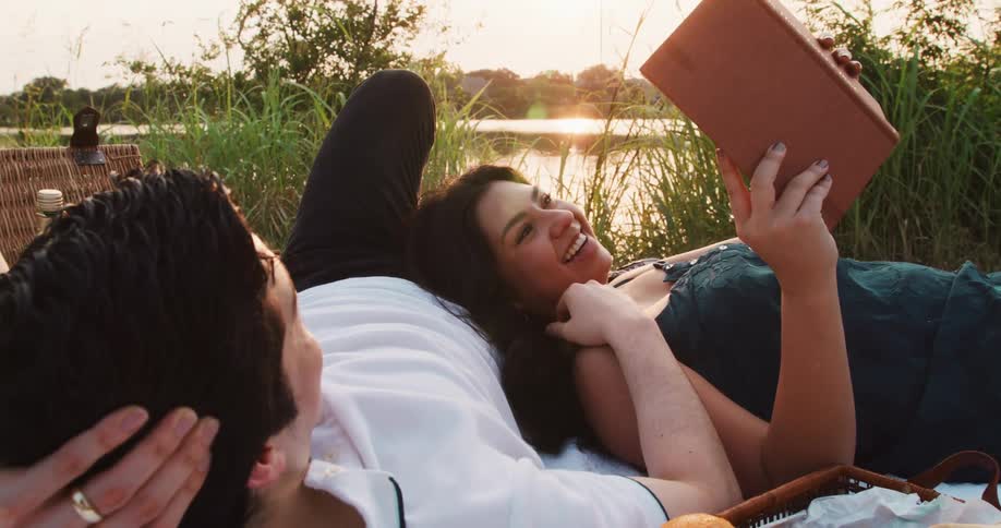 A woman smiles and kisses her husbands hand while they lie next to a lake