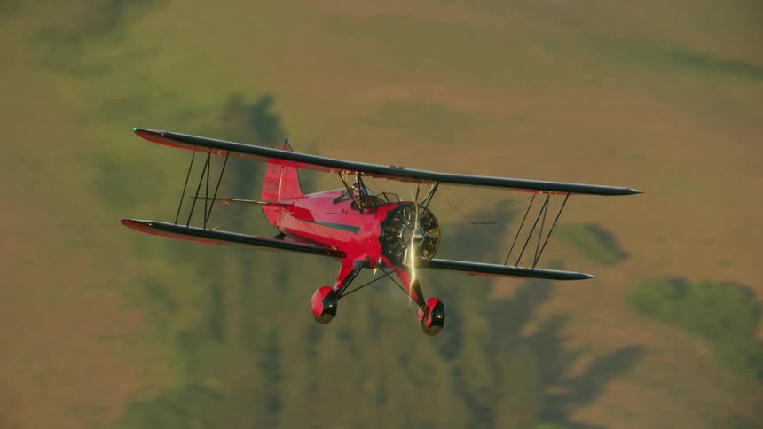 A pilot operates a small red biplane over a rural mountains and hills