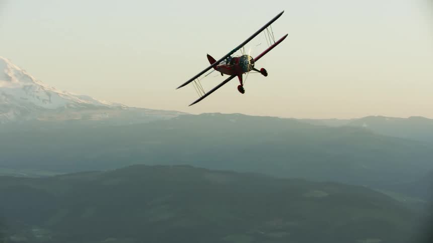 A biplane flying in the air by making s-turns in front of a high alpine peak