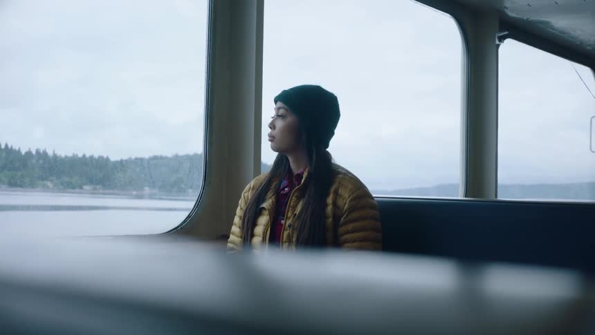 A woman sitting on a bench seat looking out the large windows of a ferry