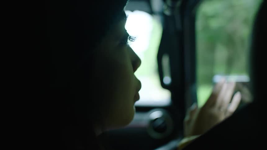 A woman is sitting in an automobile with her hand on the window as she looks outside