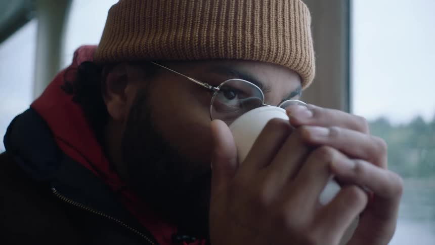 A man holds his coffee cup in both hands to warm them while looking out the ferry window