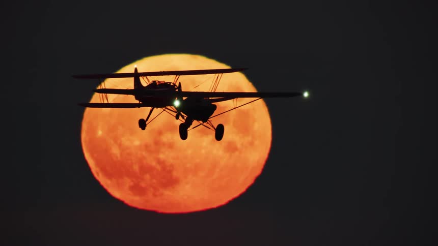 Two small planes flying in tandem are silhouetted against the red moon in the night sky
