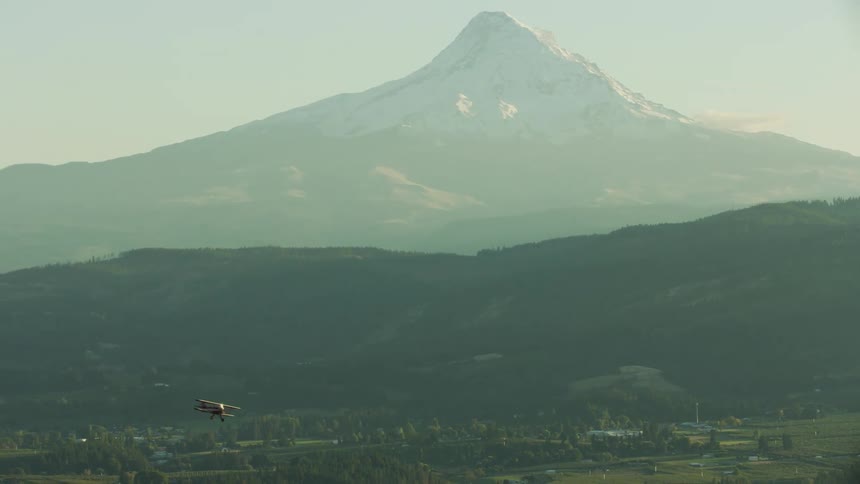 Aerial view of a red biplane flying over a small town near a mountain at morning