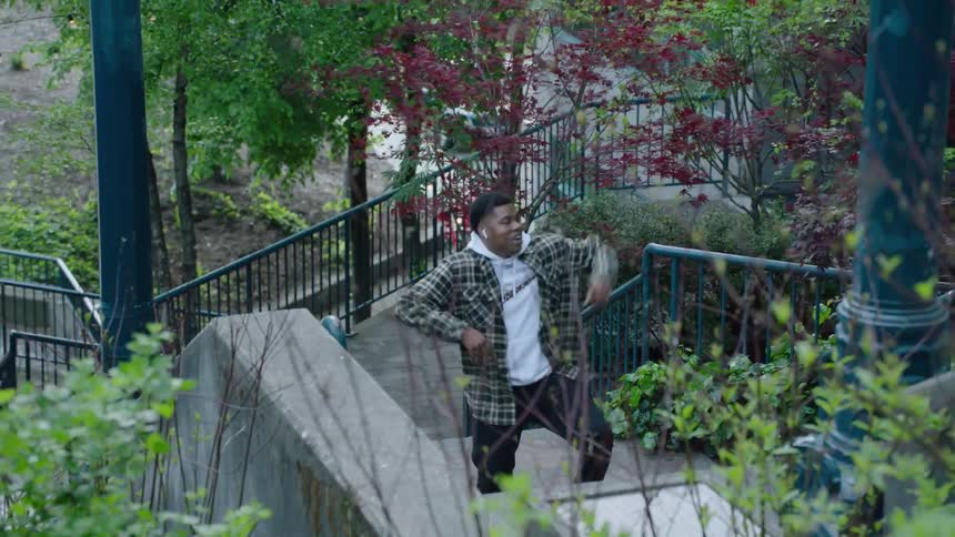 Man with earphones white hoodie green jacket and black pants daces going up the stairs in the park