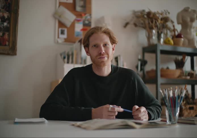 A man is sitting at a table with an open book in front of him and he is holding a pen as he looks ahead