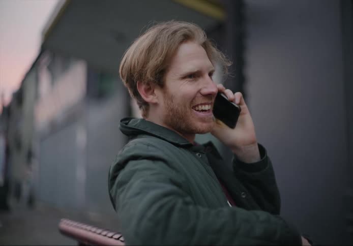 A young white man is sitting on a bench by his dog and talking on the phone