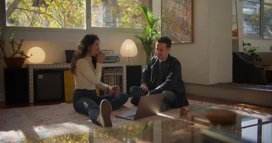 Young couple sitting in the living room floor talk while browsing on a laptop