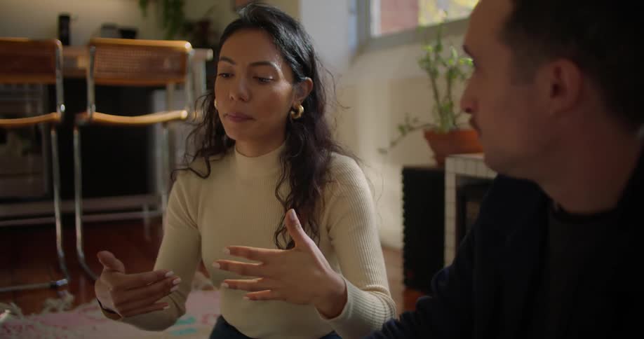 A woman and a man are sitting on a living room floor and she is moving her hands as she explains something to him
