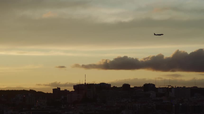 An airplane flying across the sky above the silhouette of buildings and antennas