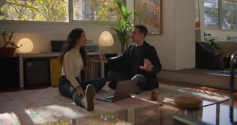 Young couple sitting in the living room floor talk while browsing on a laptop