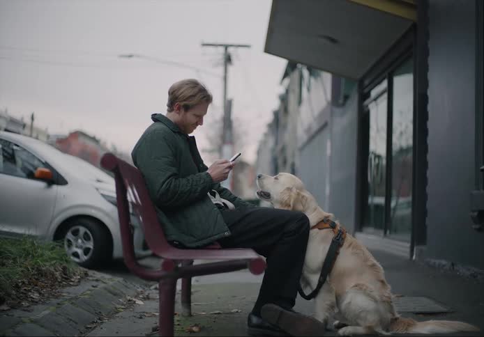 A man sitting on a sidewalk bench petting his dog and scrolling on his phone