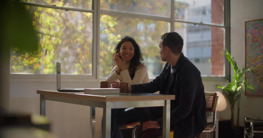 A man and a woman are sitting at a table and he is speaking as she has her hand on her laptop computer and she laughs