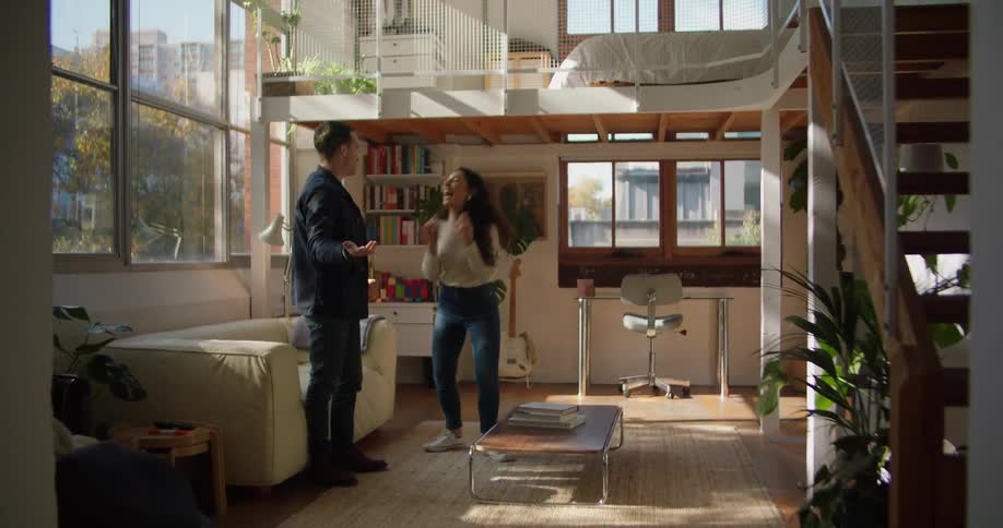 Man sets down coffee table and woman hugs him before they sit on couch near window with view of apartments