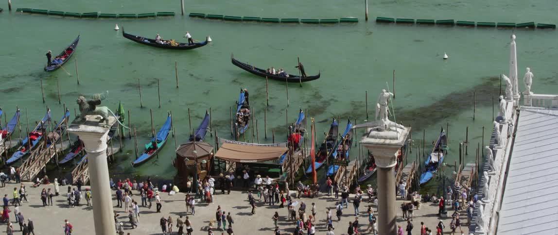 Crowd moves along shore near towers and building topped with statues as boats are poled away from shore