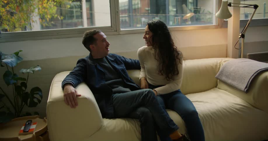 A man and woman are standing in a room and they hug and he lifts her off her feet and they fall down on to a couch and laugh