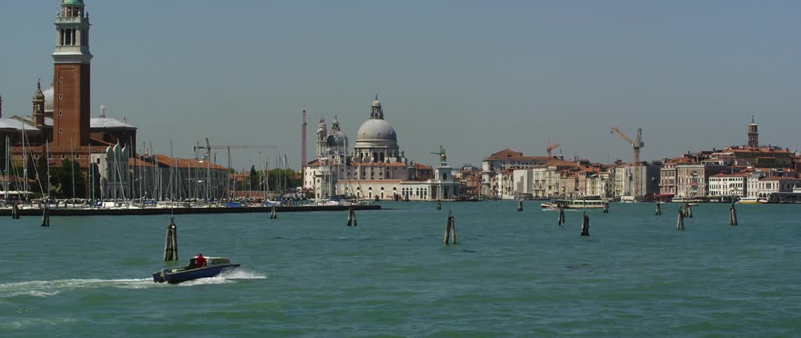 A person steers a motorboat along a waterway on the coast of a city during the day