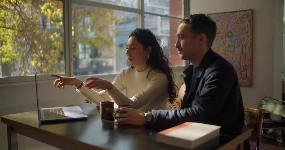 Young couple sitting at a table in the living room talk while looking at a laptop