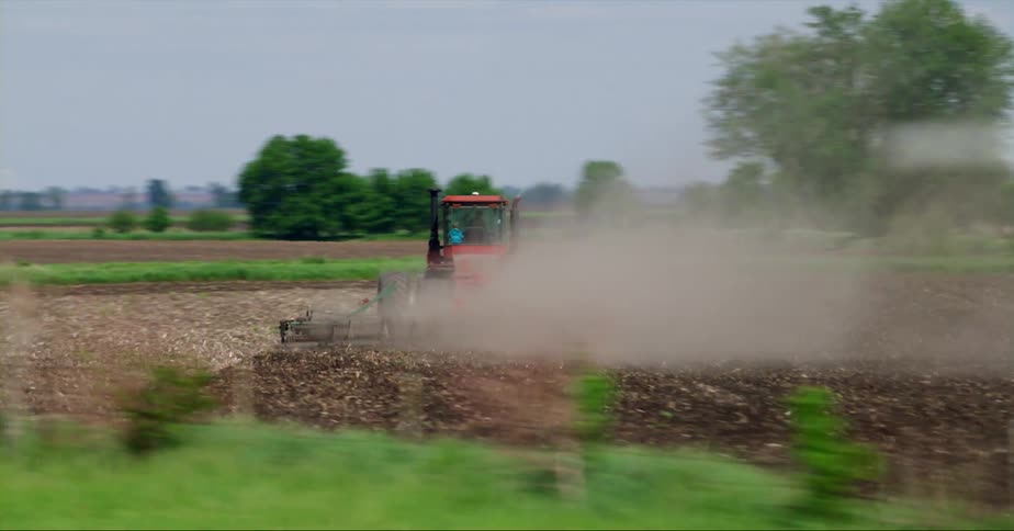 Red tractor tiling a field on a farm close to the road seen from a moving vehicle