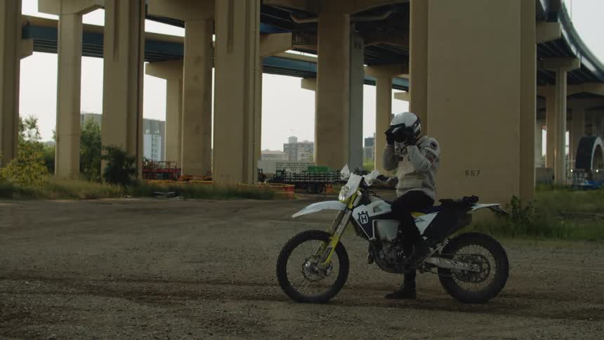A rider sitting on their dirt bike under the elevated highway and tracks adjusting their goggles