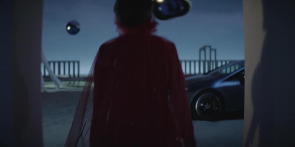 A woman in a red dress walks towards a car parked on a sandy ground