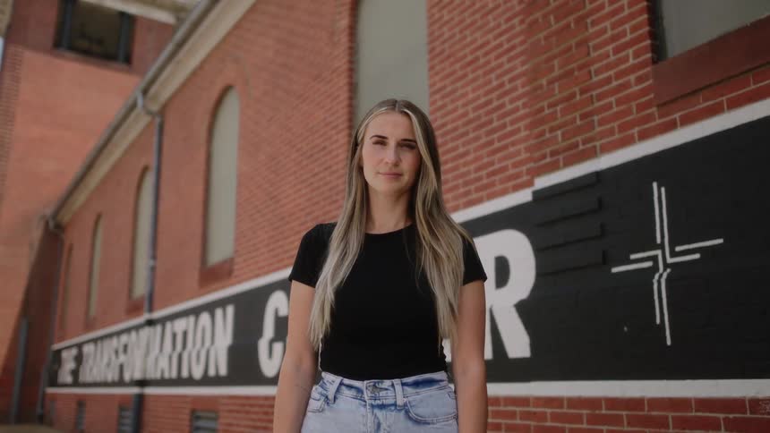 Young blonde woman standing in front of a red brick wall looking at the camera