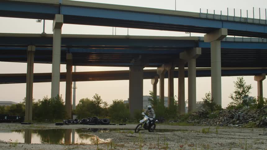 A cyclist riding a motorbike below an overpass makes a sharp turn in the dirt near a water puddle
