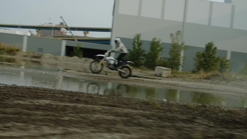 Motorcyclist pops a wheelie and rides through puddle in empty lot near bridge