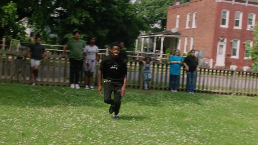 Black teenager kicks a soccer ball then runs while others lean against a fence and watch