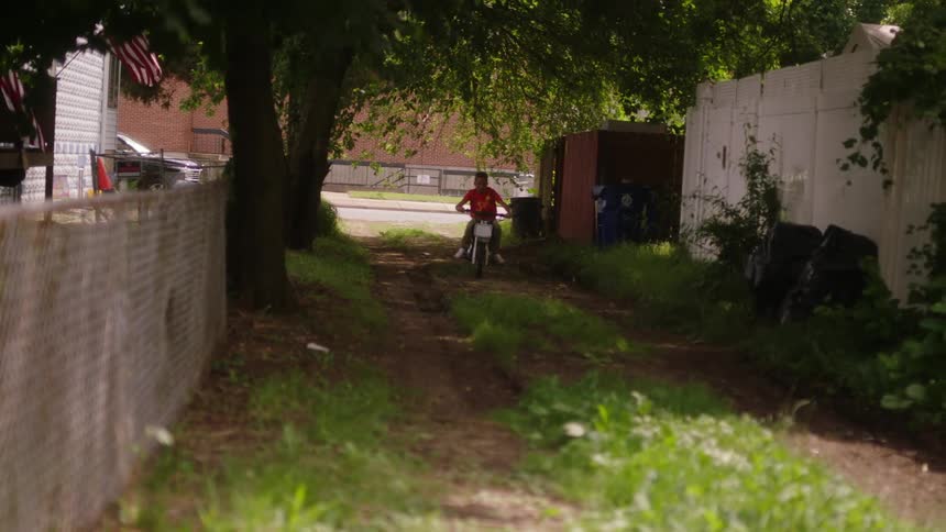 Youth rides small motorcycle along dirt alley between houses
